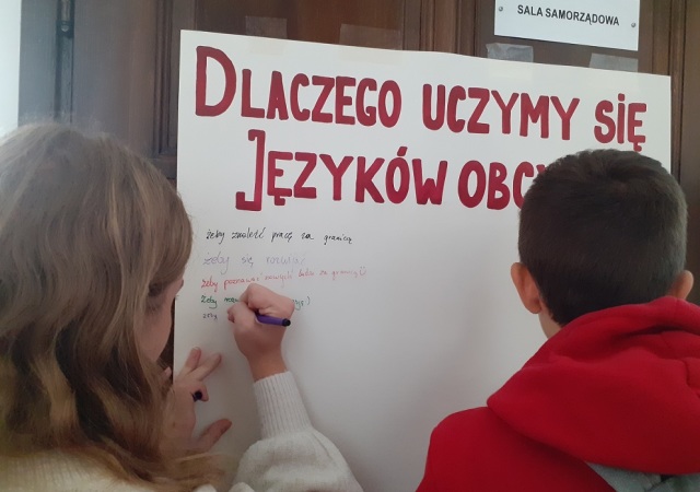 Uczennica wpisuje odpowiedź na pytanie: Dlaczego uczymy się języków obcych? które umieszczone jest na dużym arkuszu papieru. 